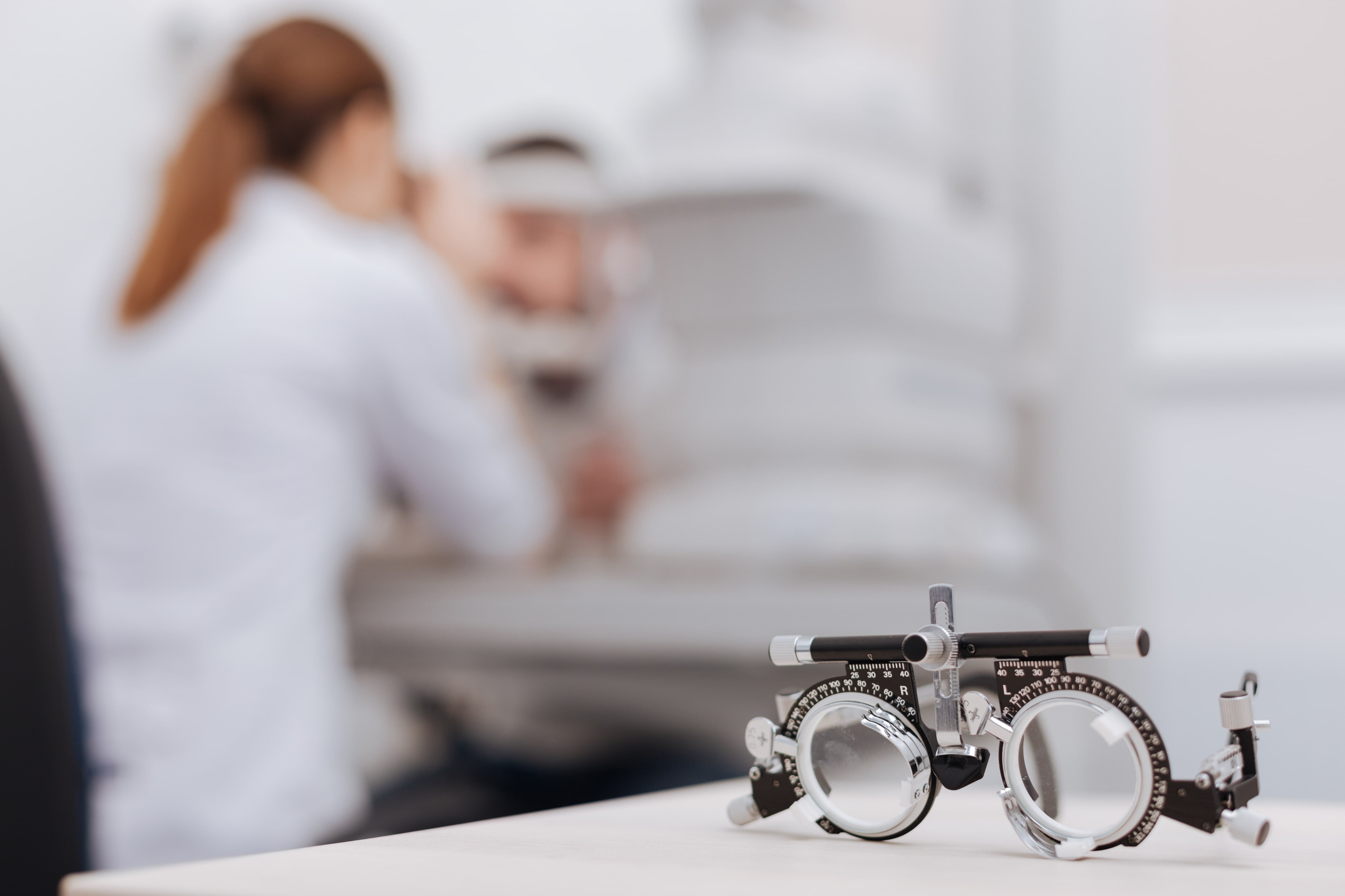 Im Hintergrund führt eine Augenärztin eine Untersuchung durch. Im Vordergrund liegt eine Augenarztbrille